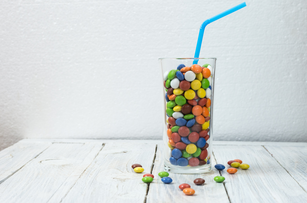 A water glass filled with sweet colorful candy with a blue straw sticking out of it and candy pieces scattered on the table around the glass.